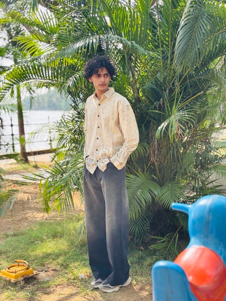 Young man with curly hair standing outdoors beside lush palm plants near a waterfront, wearing a beige patterned shirt, dark wide-leg trousers, and sneakers, looking to the side with hands in pockets.